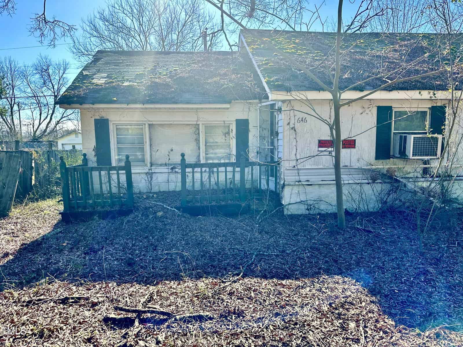 A small, weathered white house with a front porch and a window air conditioning unit, partially obscured by overgrown vegetation and a wooden fence, under a clear blue sky.