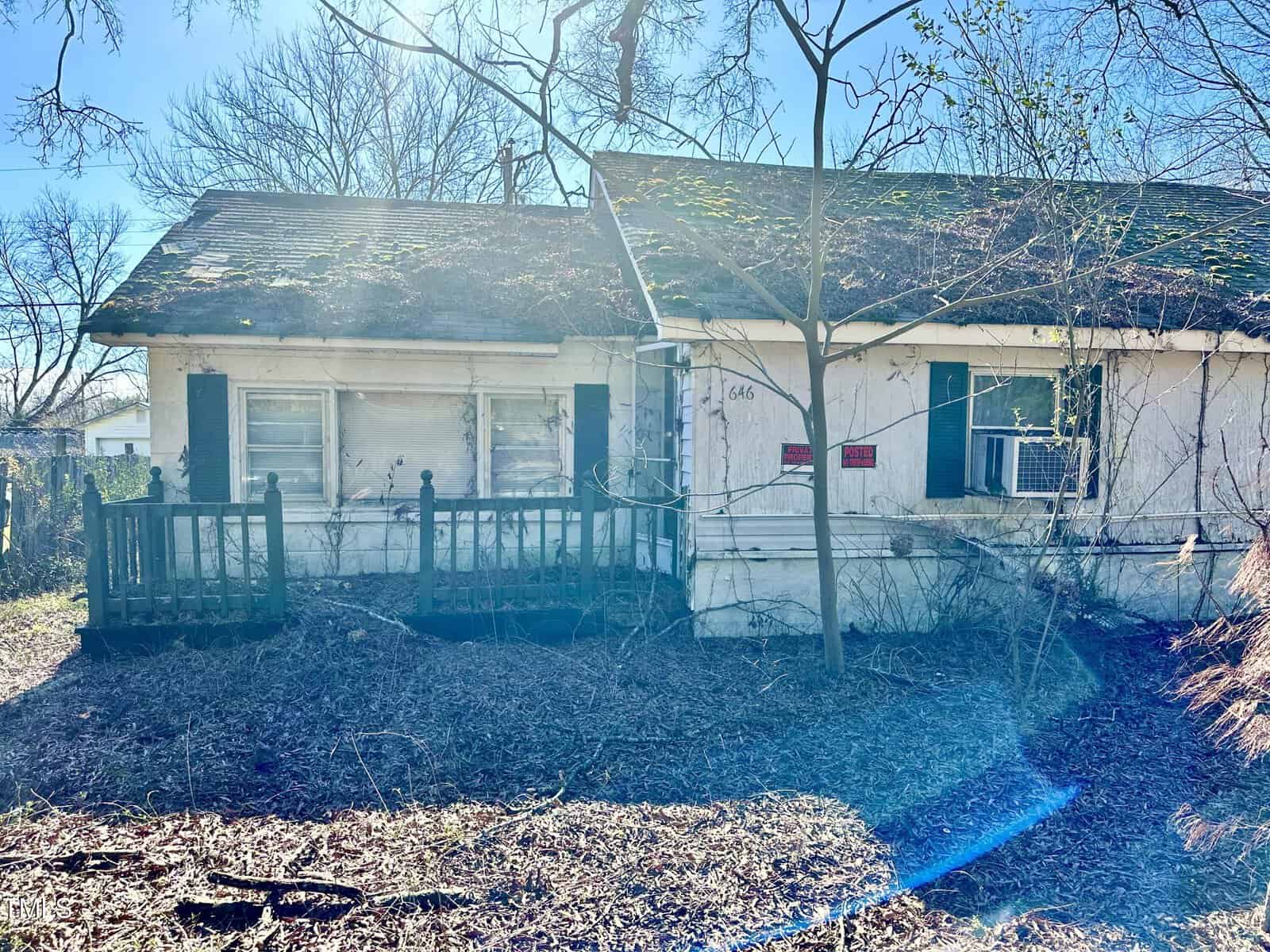 A single-story house with a white exterior, a small porch, and a bare tree in the front yard under bright sunlight with visible lens flare.