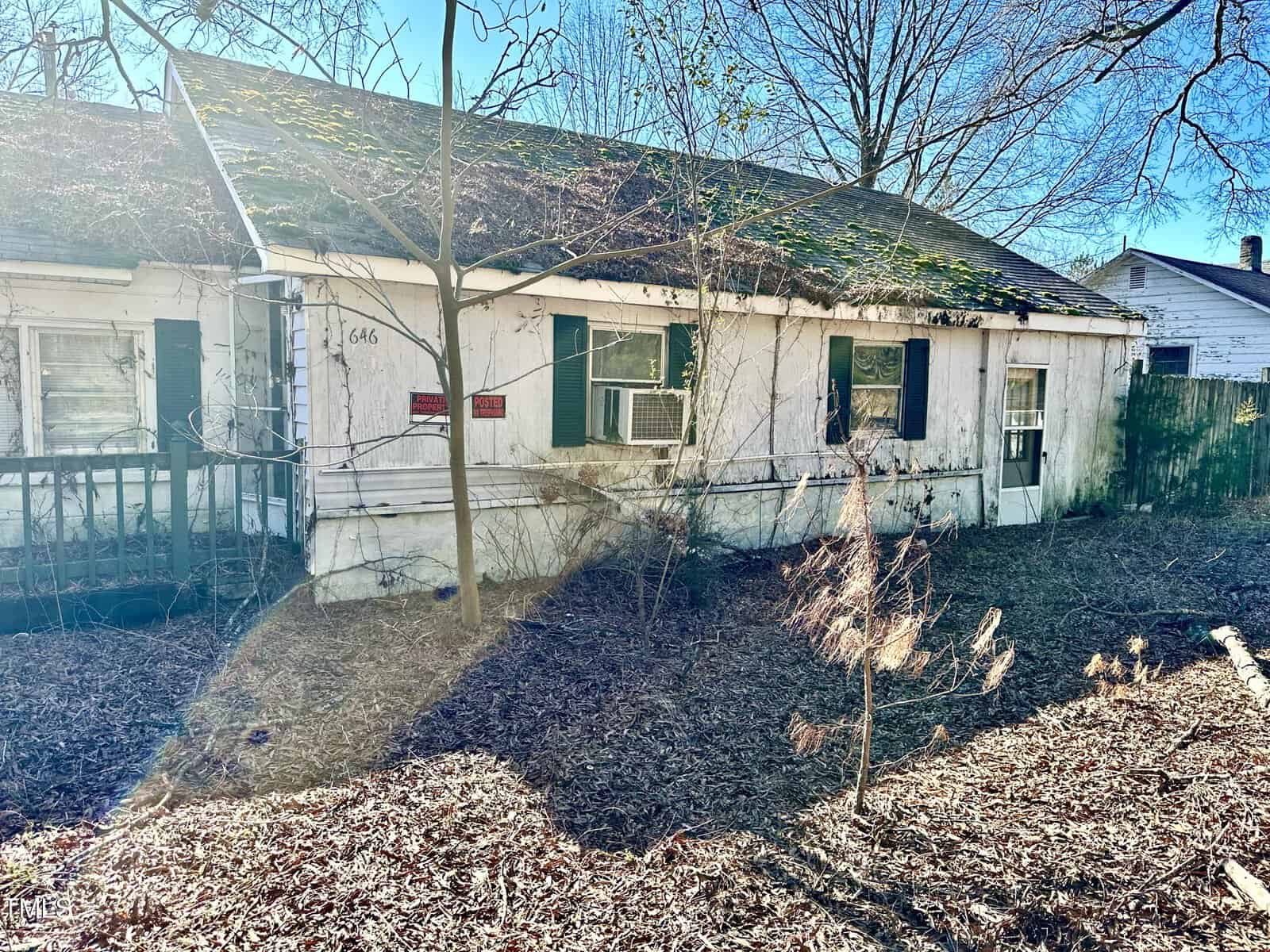 A dilapidated single-story house with peeling white paint, boarded-up windows, and an overgrown yard under a clear blue sky.