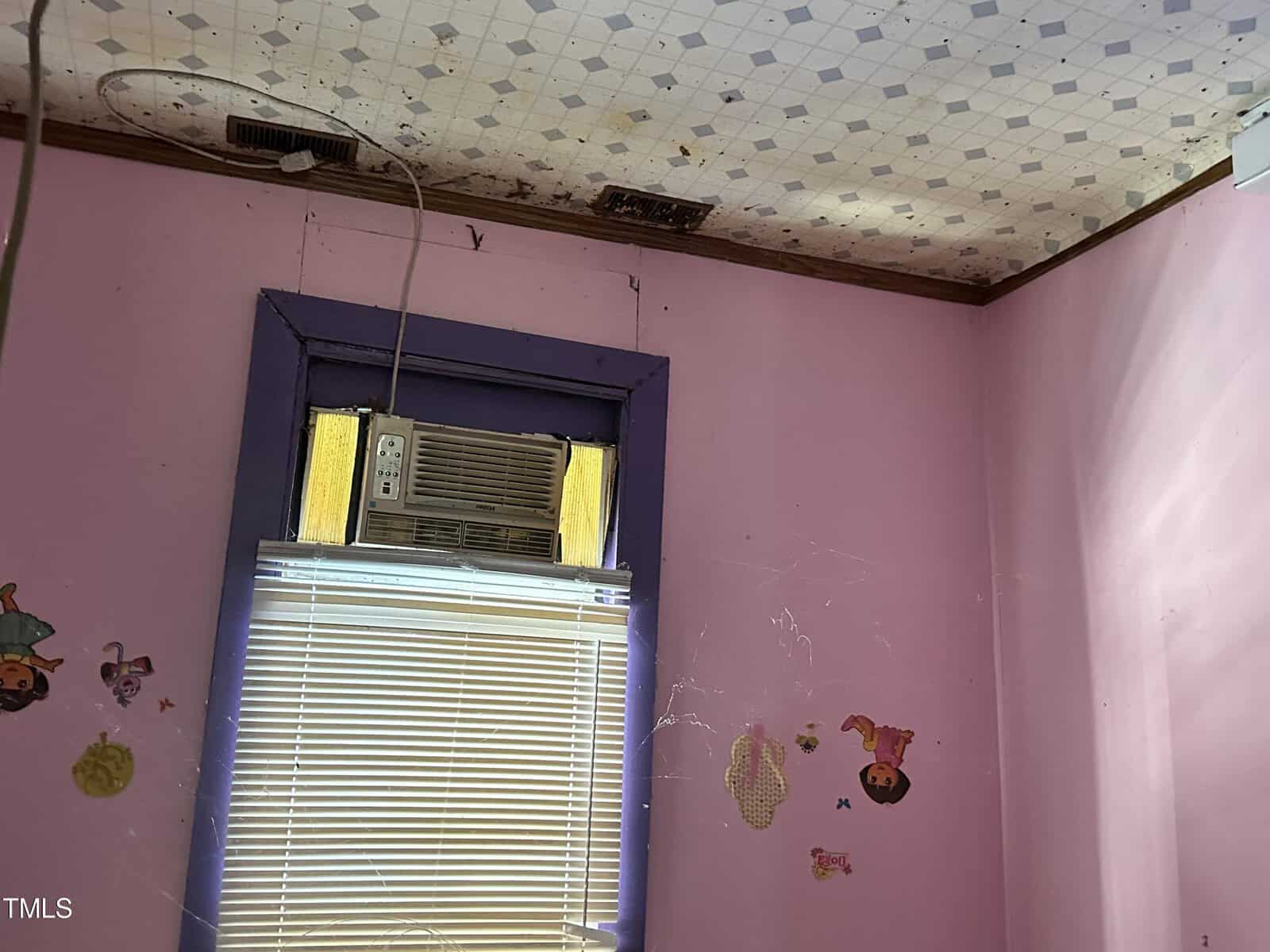 An old, neglected room with purple walls, decorative stickers, a window-mounted air conditioner, and a ceiling vent with visible dust and cobwebs.