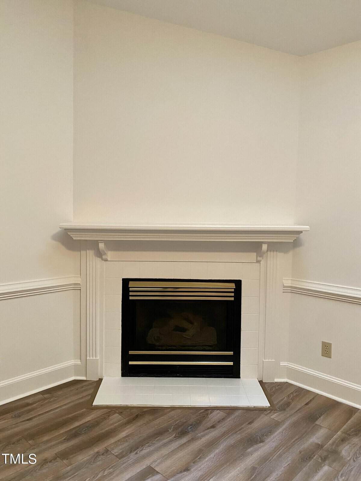 A clean, white fireplace with a closed glass door and imitation logs, set against a white wall with wainscoting, above herringbone-patterned wooden flooring.