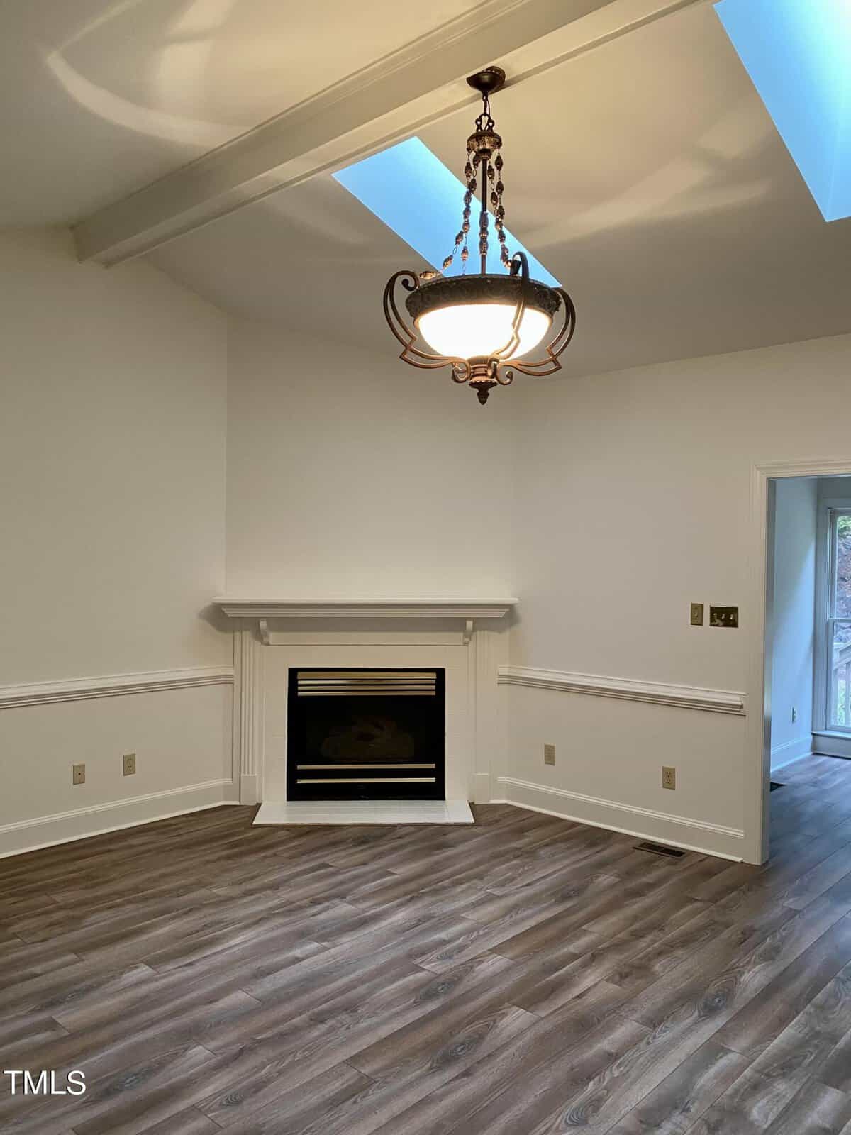 An empty room featuring a white fireplace, wood flooring, and a hanging light fixture with a vaulted ceiling.