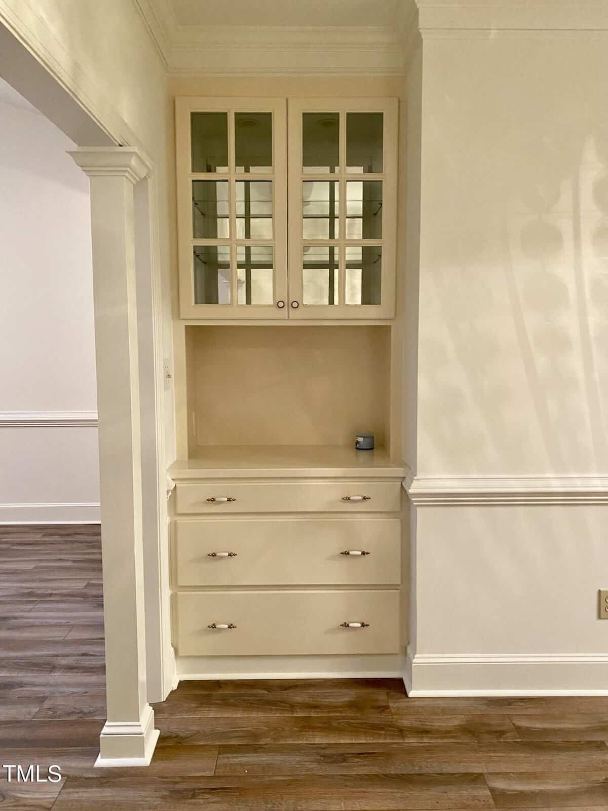 A built-in wall cabinet with glass-paneled upper doors and three lower drawers in a room with beige walls and wooden flooring.