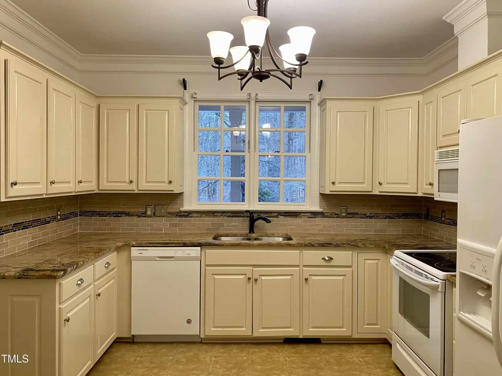 A well-lit kitchen with cream-colored cabinets, black countertops, and white appliances, featuring a window above the sink.