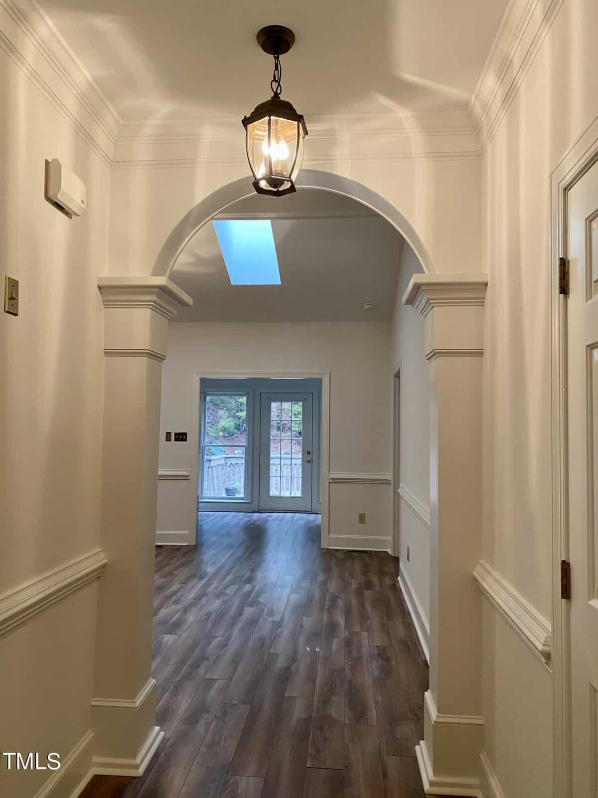 An interior hallway in a residential home with an arched doorway, paneled walls, and hardwood flooring, leading to a room with a glass door.