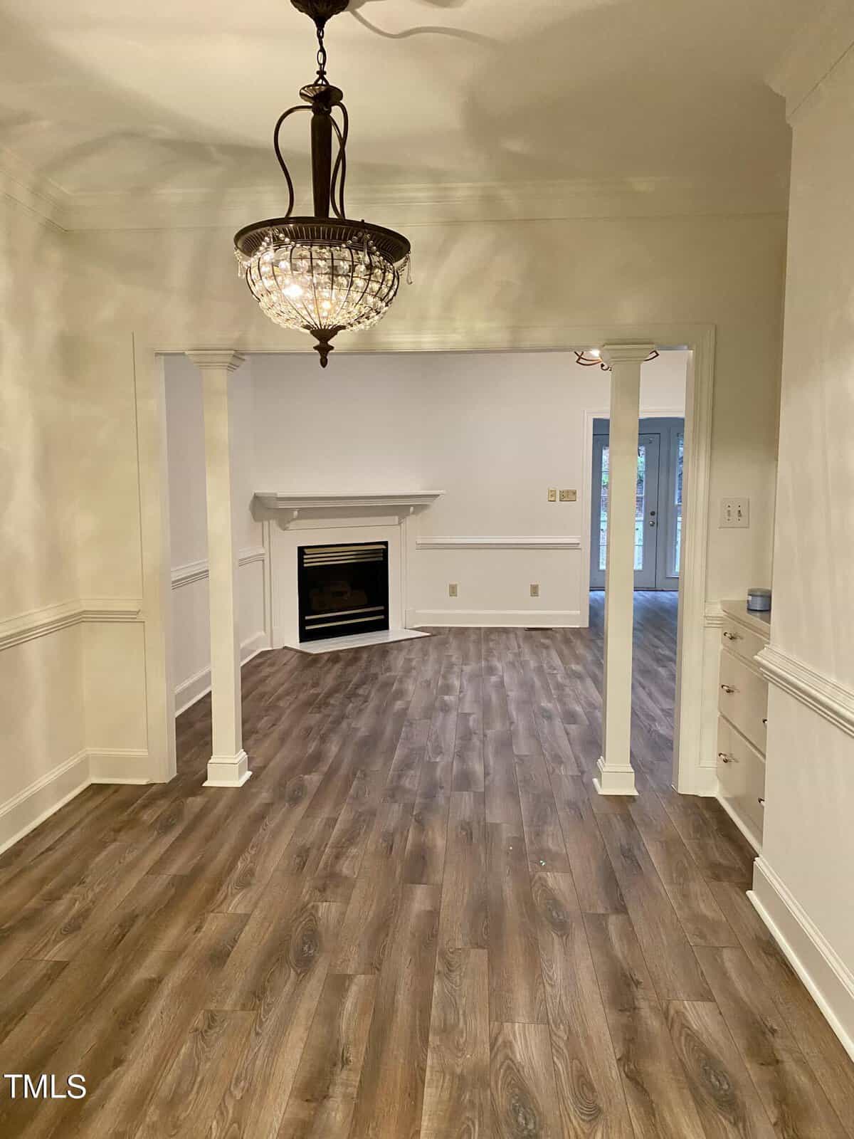An empty room featuring wood flooring, white walls with wainscoting, a fireplace, and a hanging crystal chandelier.