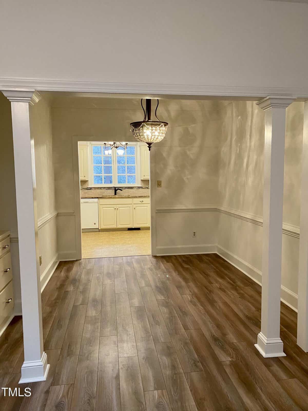 An interior view of a home showcasing an empty room with hardwood flooring, featuring white columns and half-wall that lead to a kitchen with white cabinetry and a window. a hanging light fixture is visible above.