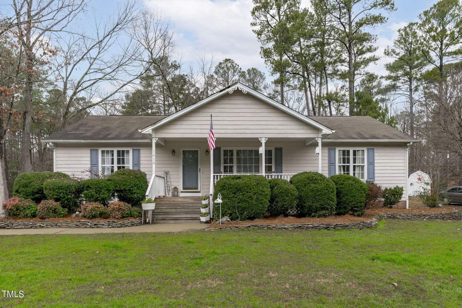 Nestled on 1410 W B Street, this single-story house in Butner showcases a charming front porch surrounded by trimmed bushes and a lush lawn. An American flag proudly hangs by the entrance, with tall trees and a partly cloudy sky as its serene backdrop.
