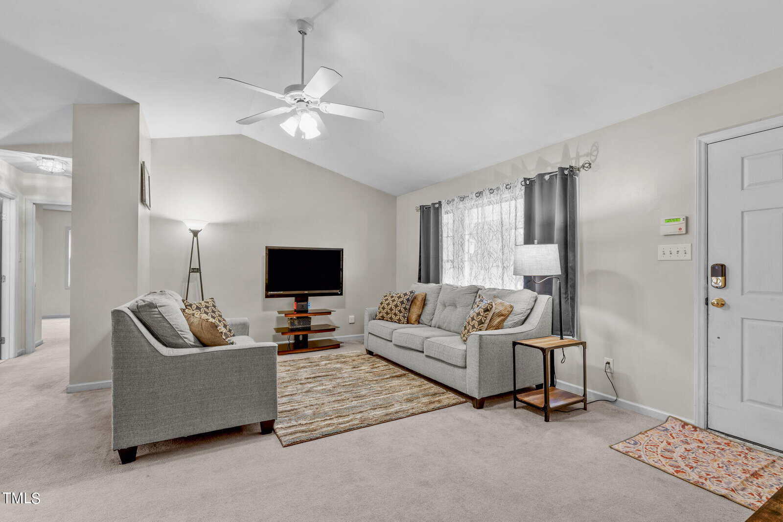 A cozy living room with two gray sofas facing each other, a TV on a stand, and a floor lamp in the corner. A patterned brown rug is on the carpeted floor. The room is lit by natural light through a curtained window and a ceiling fan light.