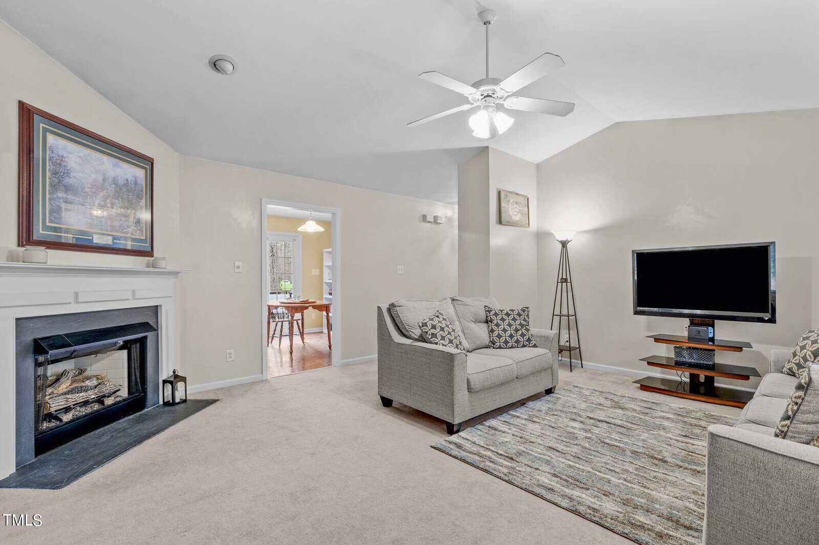 A cozy living room with beige walls and carpet, featuring a fireplace on the left, a ceiling fan, two gray sofas, a flat-screen TV on a stand, and a rug. There's a doorway leading to a room with a table and chairs.