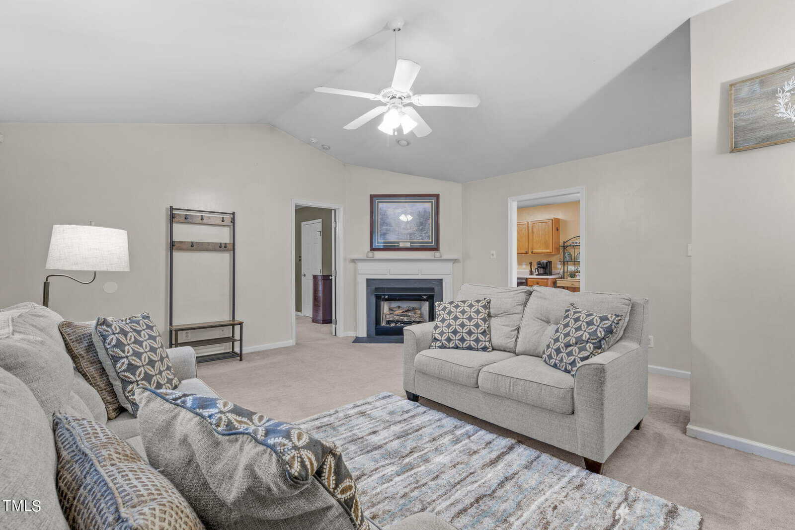 A spacious living room with a vaulted ceiling and a ceiling fan. There's a fireplace with a framed picture above it, surrounded by two beige sofas with patterned cushions. A lamp and a patterned rug add to the cozy ambiance. An open doorway leads to a kitchen.