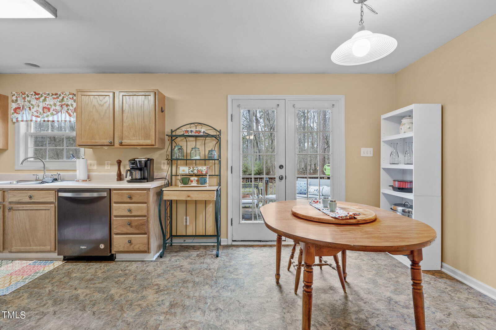 A cozy kitchen features wooden cabinets, a countertop with a sink, and a coffee maker. A round wooden table has a plate of food. French doors lead outside, next to a white bookshelf and wall-mounted light. A colorful rug adds a touch of brightness.