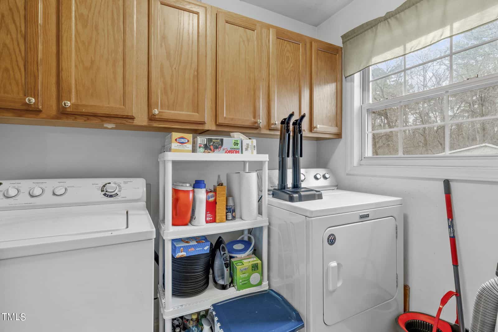 A laundry room with a washing machine and dryer, wooden cabinets, a small white shelf holding detergent and cleaning supplies, and a window with a green valance. A vacuum cleaner and a broom are nearby.