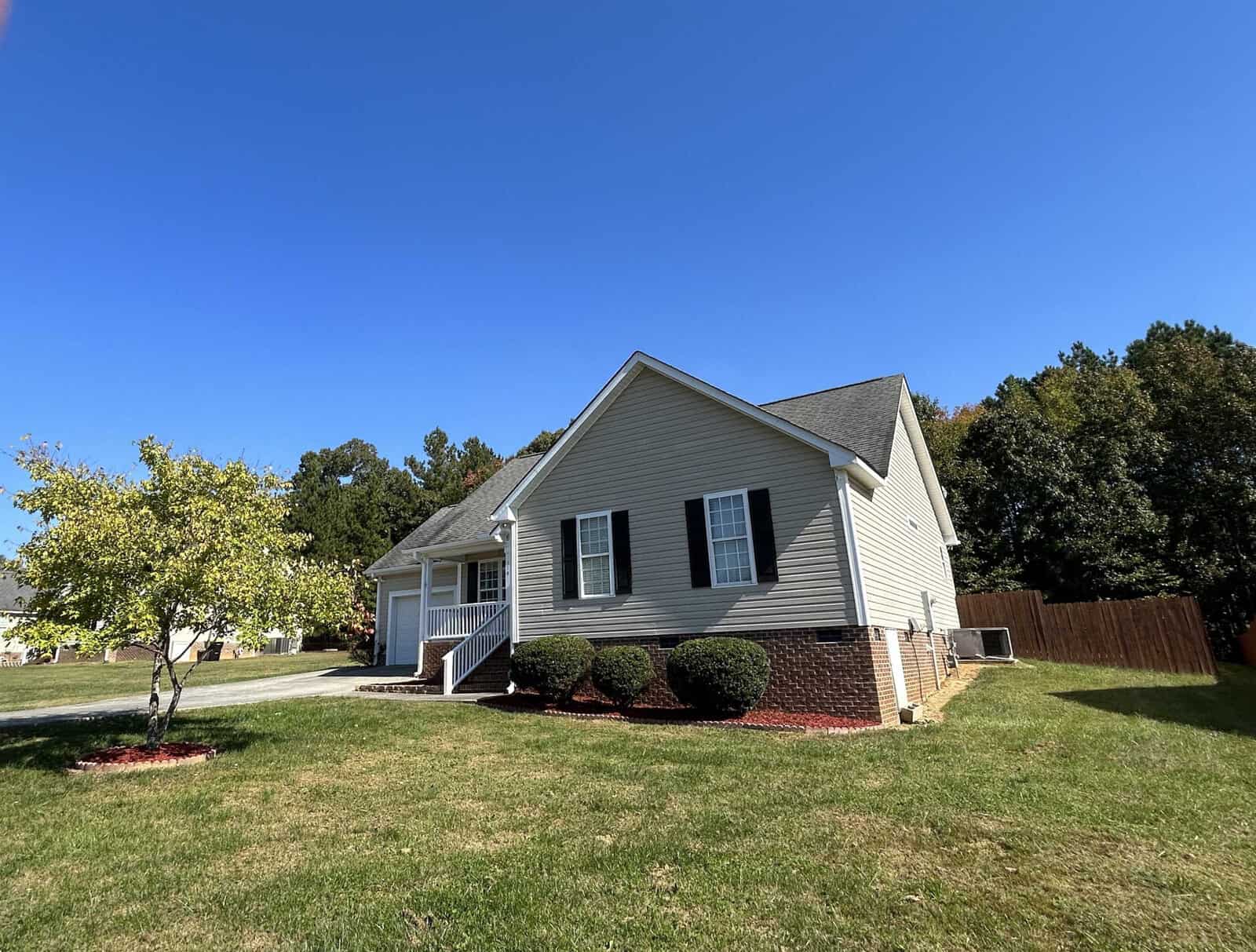 A single-story house with light gray siding and a dark shingled roof, surrounded by a green lawn and a few small trees. A sidewalk leads to the front porch under a clear blue sky. A tall wooden fence is visible in the background.