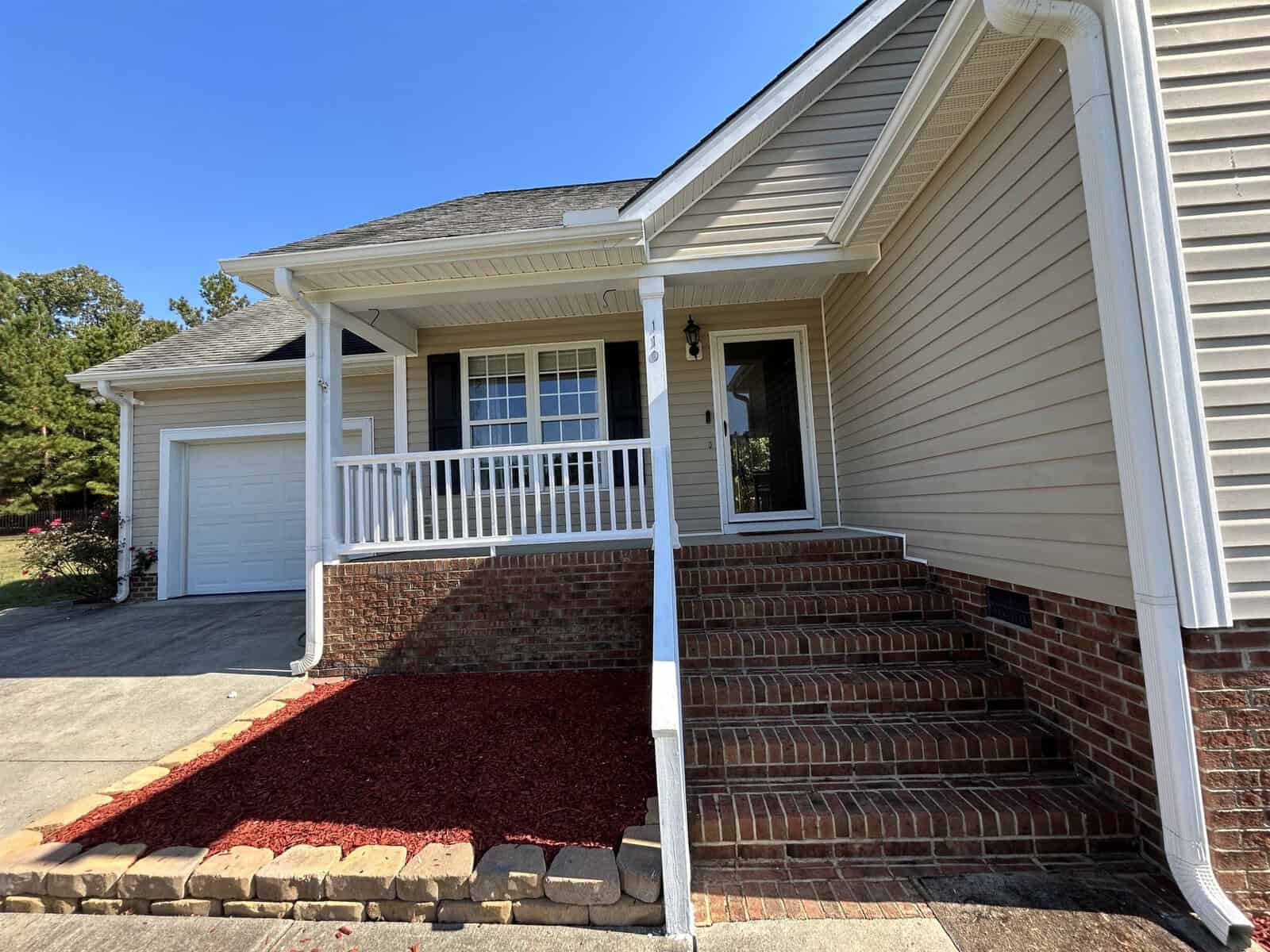A beige single-story house with white trim and a gabled roof. The front features a small porch with white railing, brick steps, and a red mulch flower bed alongside a concrete driveway leading to a garage.