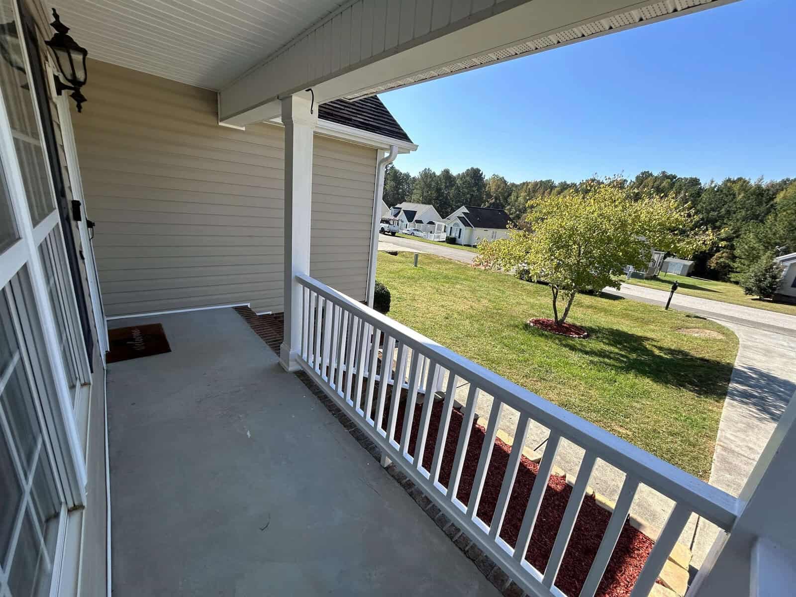 A front porch with a white railing overlooks a suburban street. The porch has a welcome mat and a light fixture next to the house's siding. In the background, there's a lawn with a tree and neighboring houses under a clear blue sky.