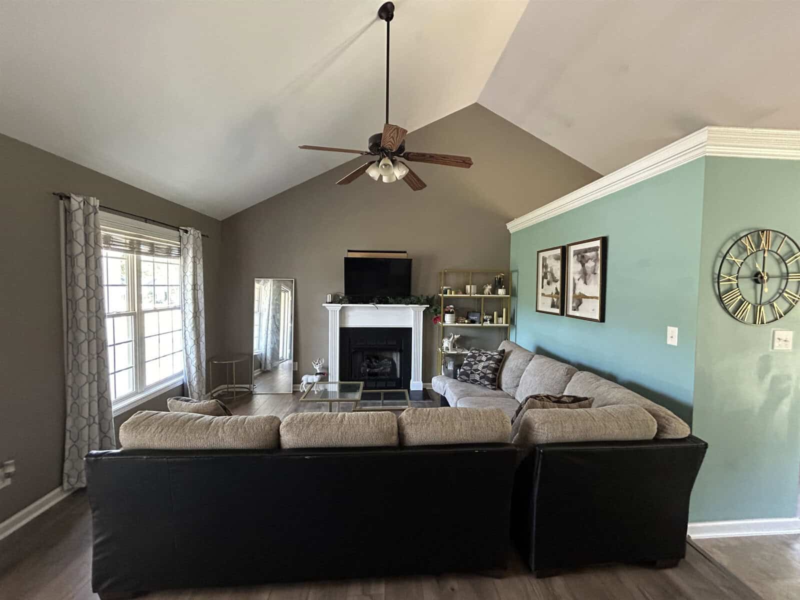 A cozy living room with a vaulted ceiling features a large sectional sofa facing a fireplace and TV. There are shelves with decorative items, a wall clock, and framed artwork. A ceiling fan is centered above, and a window with curtains allows natural light.