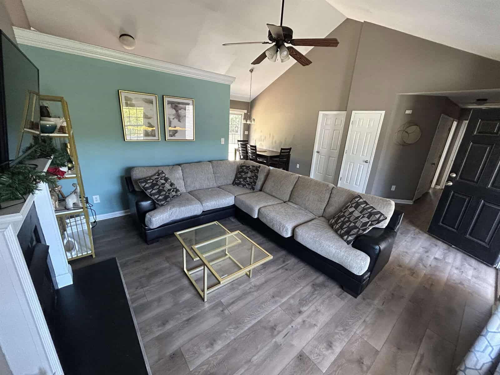 A cozy living room with a large L-shaped sofa, a glass coffee table, and a TV on the left. The walls are painted in green and beige tones. A ceiling fan hangs above, and there is a view into a dining area with a table and chairs.
