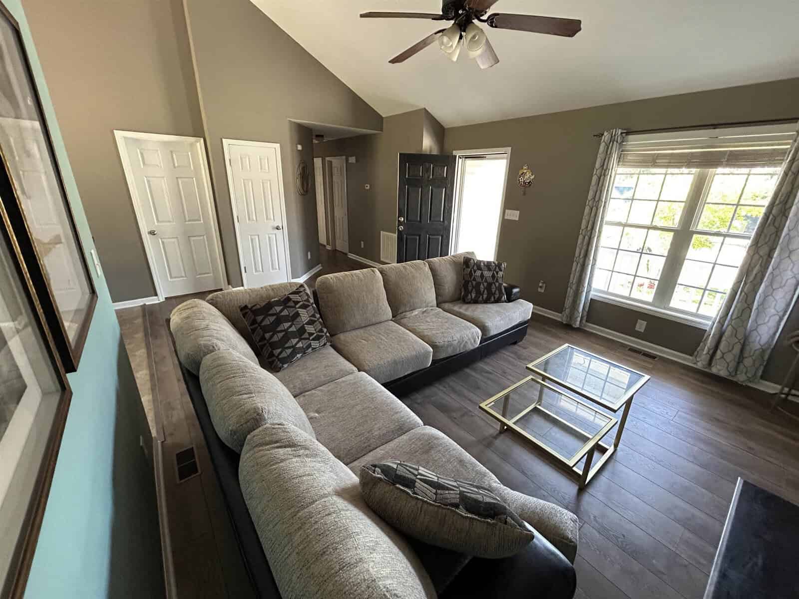 A cozy living room with a large gray sectional couch, geometric patterned pillows, a glass coffee table, and an overhead ceiling fan. Sunlight streams through large windows with patterned curtains, illuminating the wooden floor.