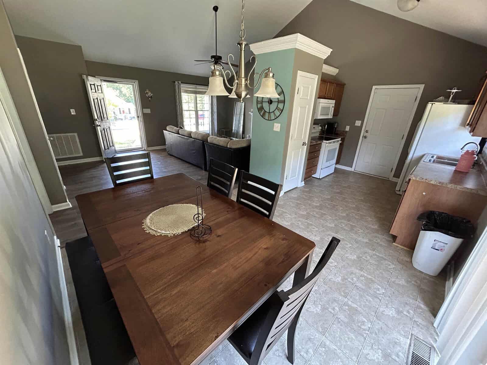 A spacious open-concept dining and living area with a wooden dining table and chairs in the foreground. The kitchen features wooden cabinets and appliances, and a sofa is visible in the living area. A light fixture hangs above the table.