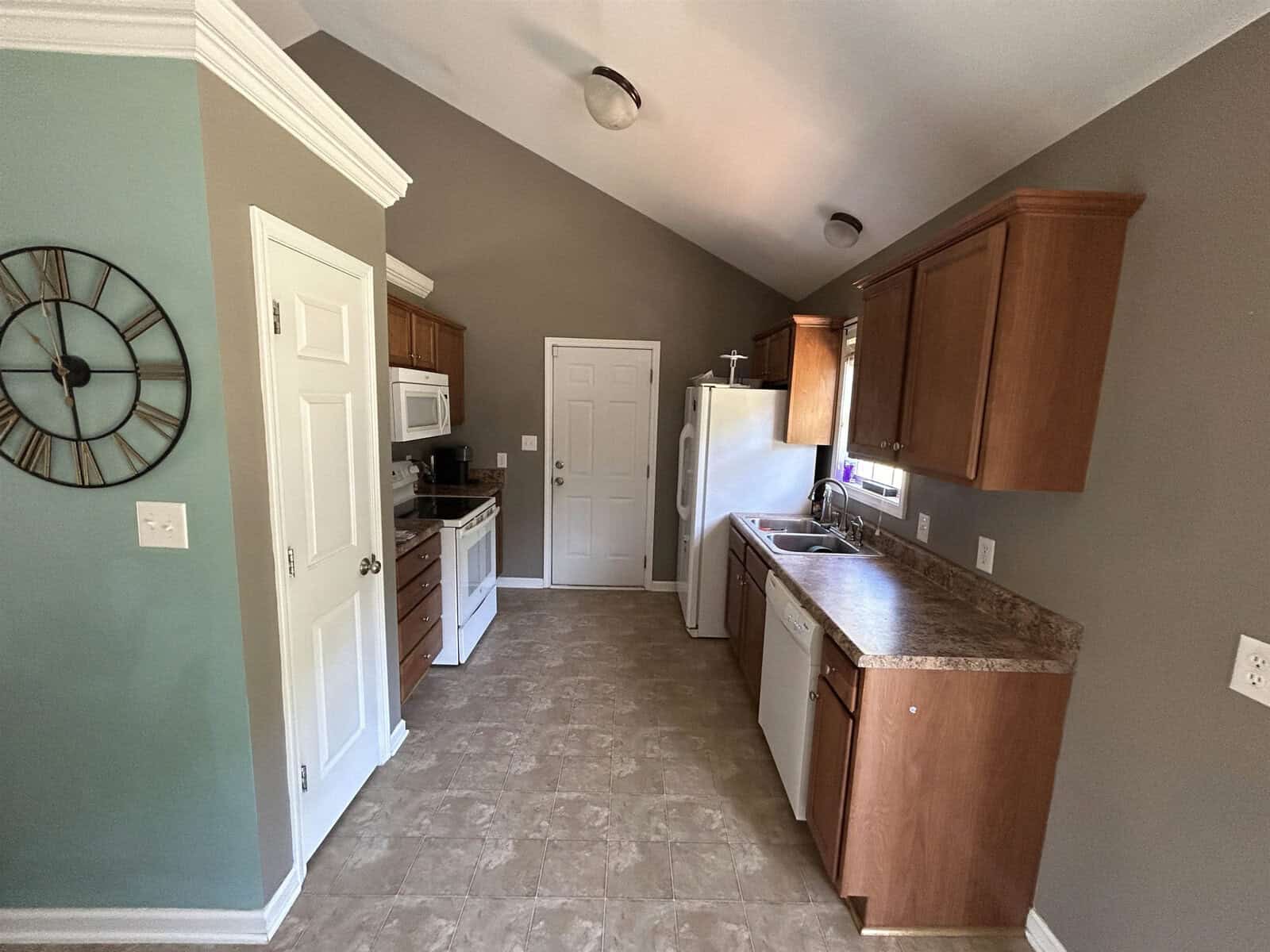 A kitchen with brown cabinets, white appliances, and a marble-patterned countertop. A wall clock is mounted on a green accent wall. The room has beige walls, tile flooring, and a door at the end.