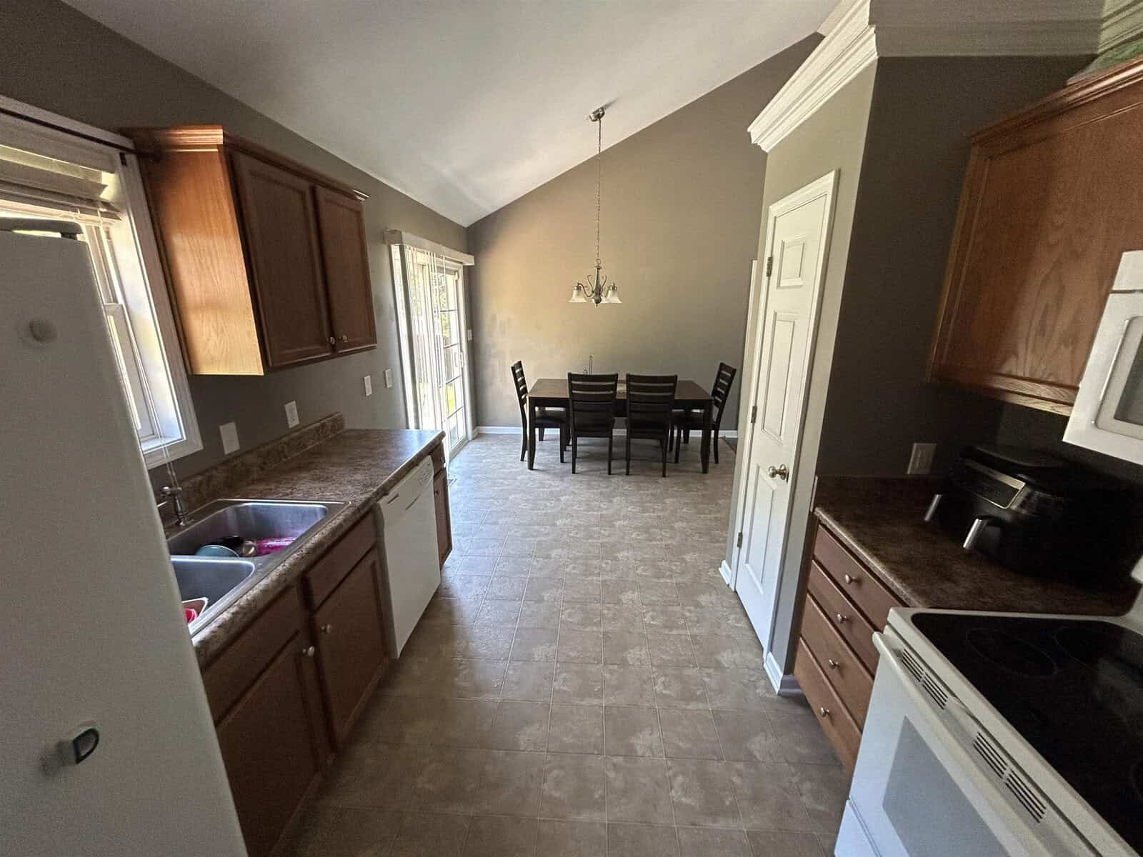 A kitchen with brown cabinets, a white refrigerator, and a white microwave. The countertop is brown, and a sink is visible. A dining area with a table and six chairs is in the background, under a chandelier and near a glass door.