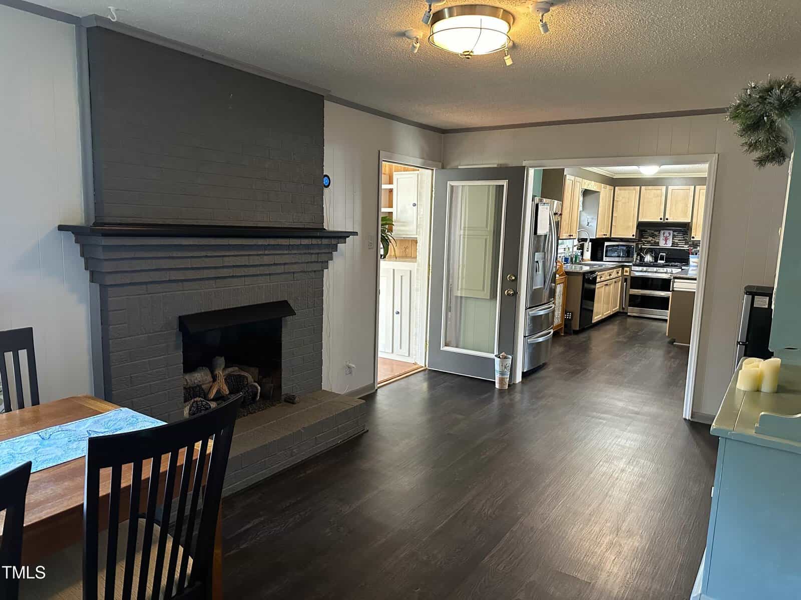 A well-lit, unfurnished room featuring a dark gray fireplace, with a view into an adjoining kitchen area that has wooden cabinets and white appliances.