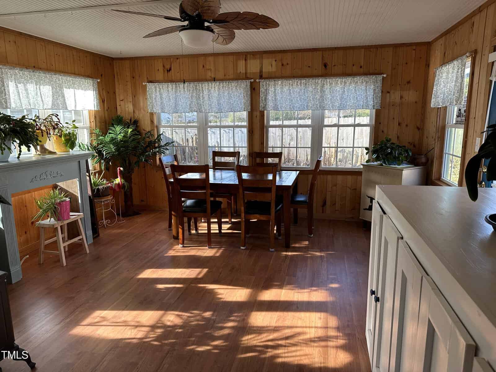 A bright and sunny dining room with a wooden table and chairs, surrounded by numerous potted plants, with sunlight casting patterns on the floor through the window blinds.