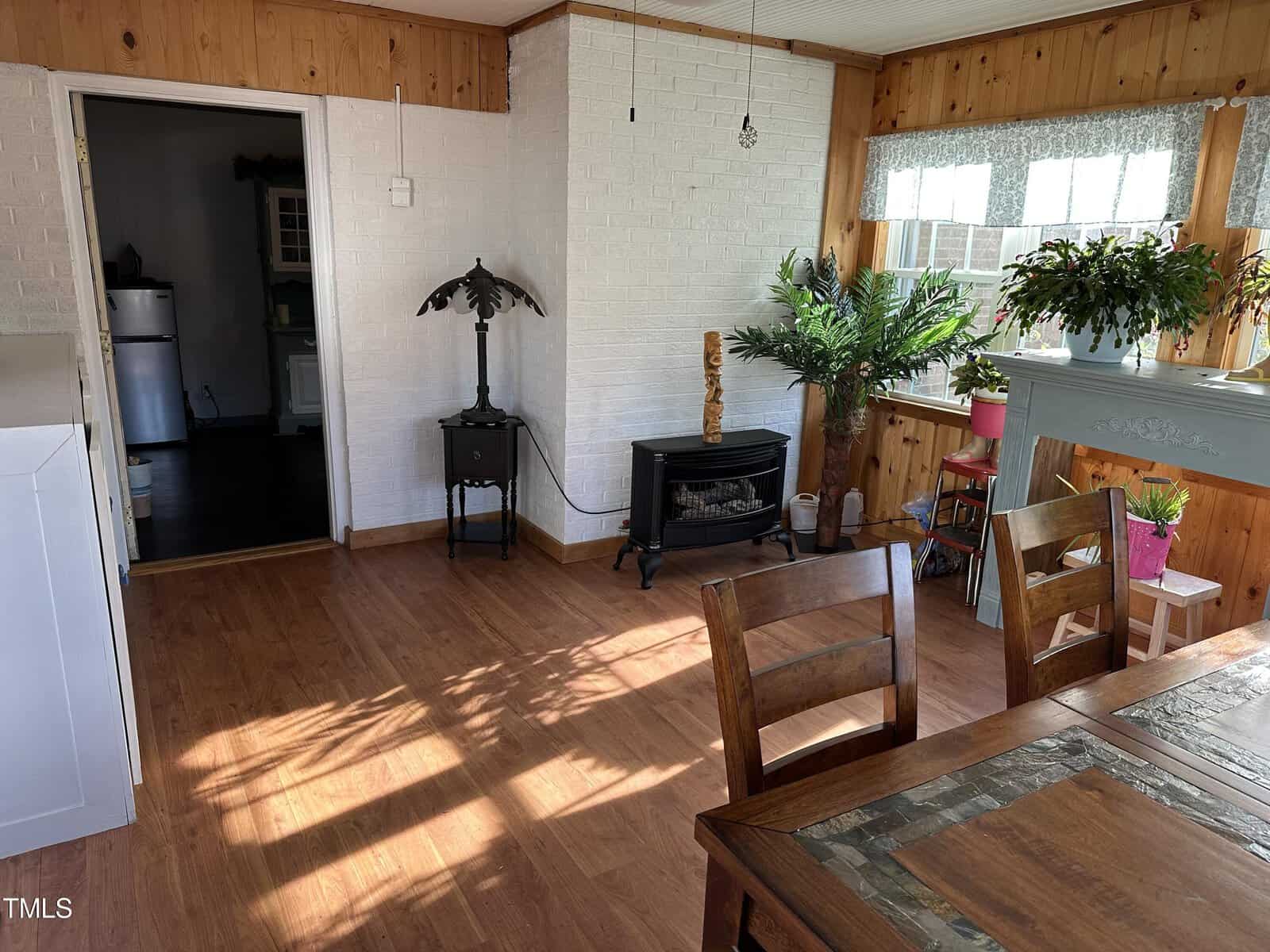 A sunlit, cozy room featuring a wooden dining table, a black wood stove, potted plants, and a mix of rustic and white-painted brick walls.