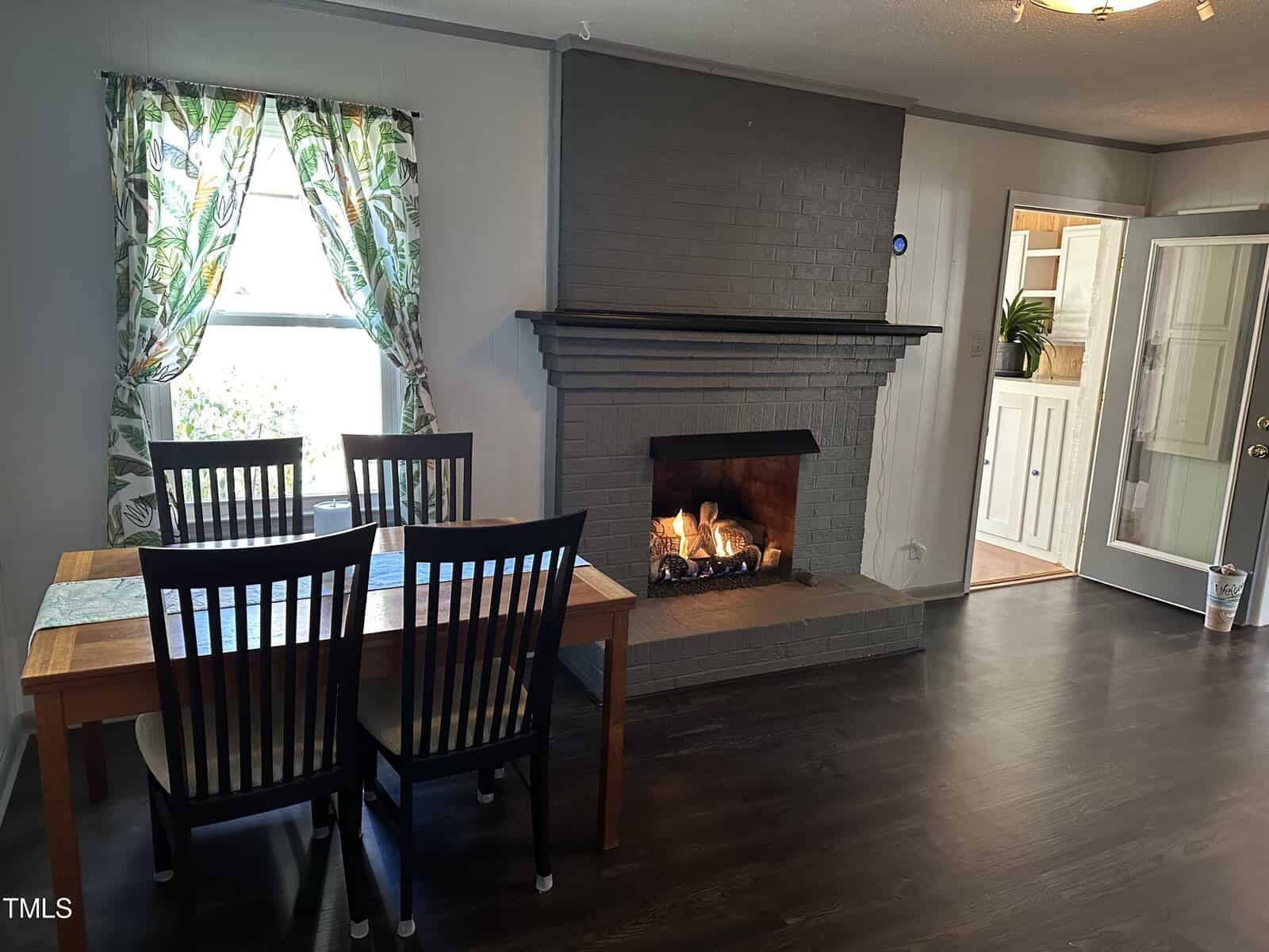 A dining area with a wooden table and four chairs adjacent to a living space with a lit fireplace, featuring patterned curtains and laminate flooring.