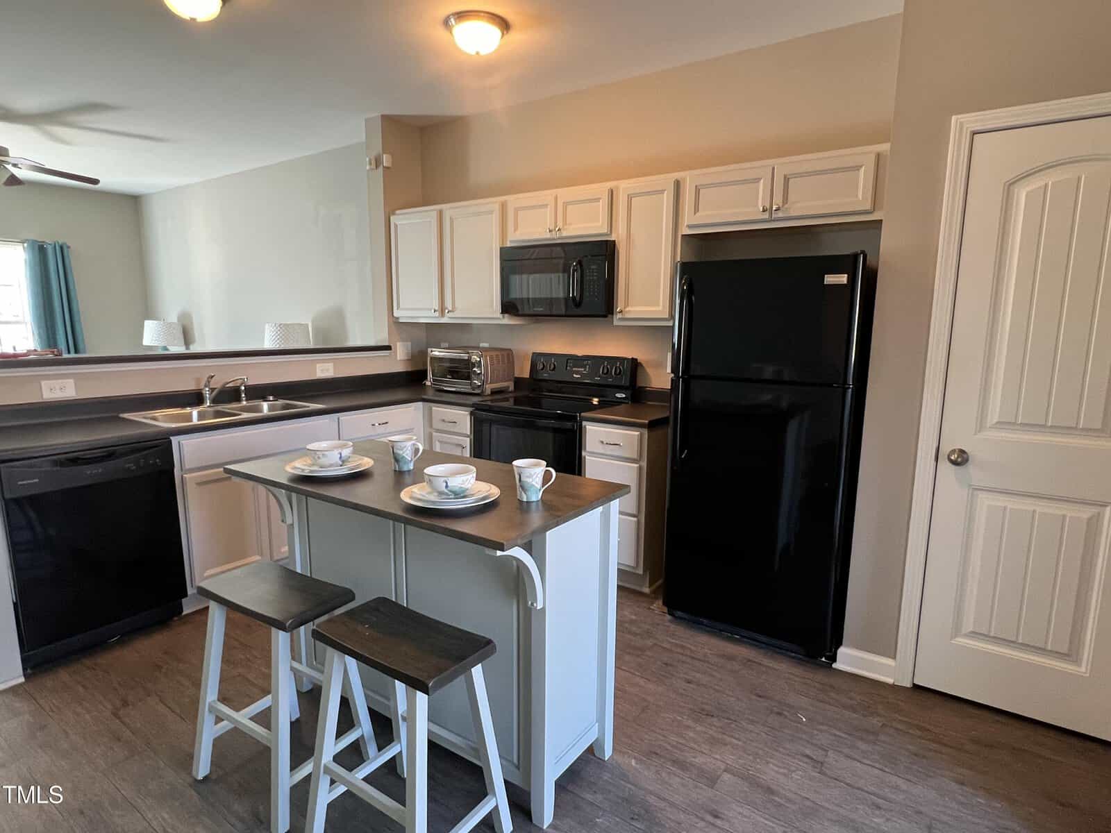 A kitchen with white cabinets and black appliances, including a refrigerator and stove. A small island with two stools is set with plates and cups. Bright lighting and a wooden floor complete the space.