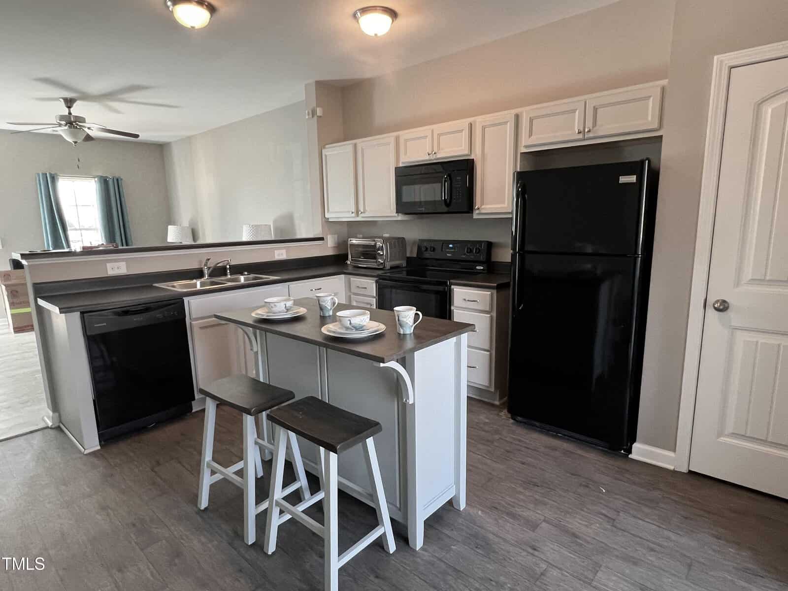 Modern kitchen with white cabinets, black appliances, and an island with two stools. The room has a ceiling fan and a partial view of a living area with a window. A door is to the right.