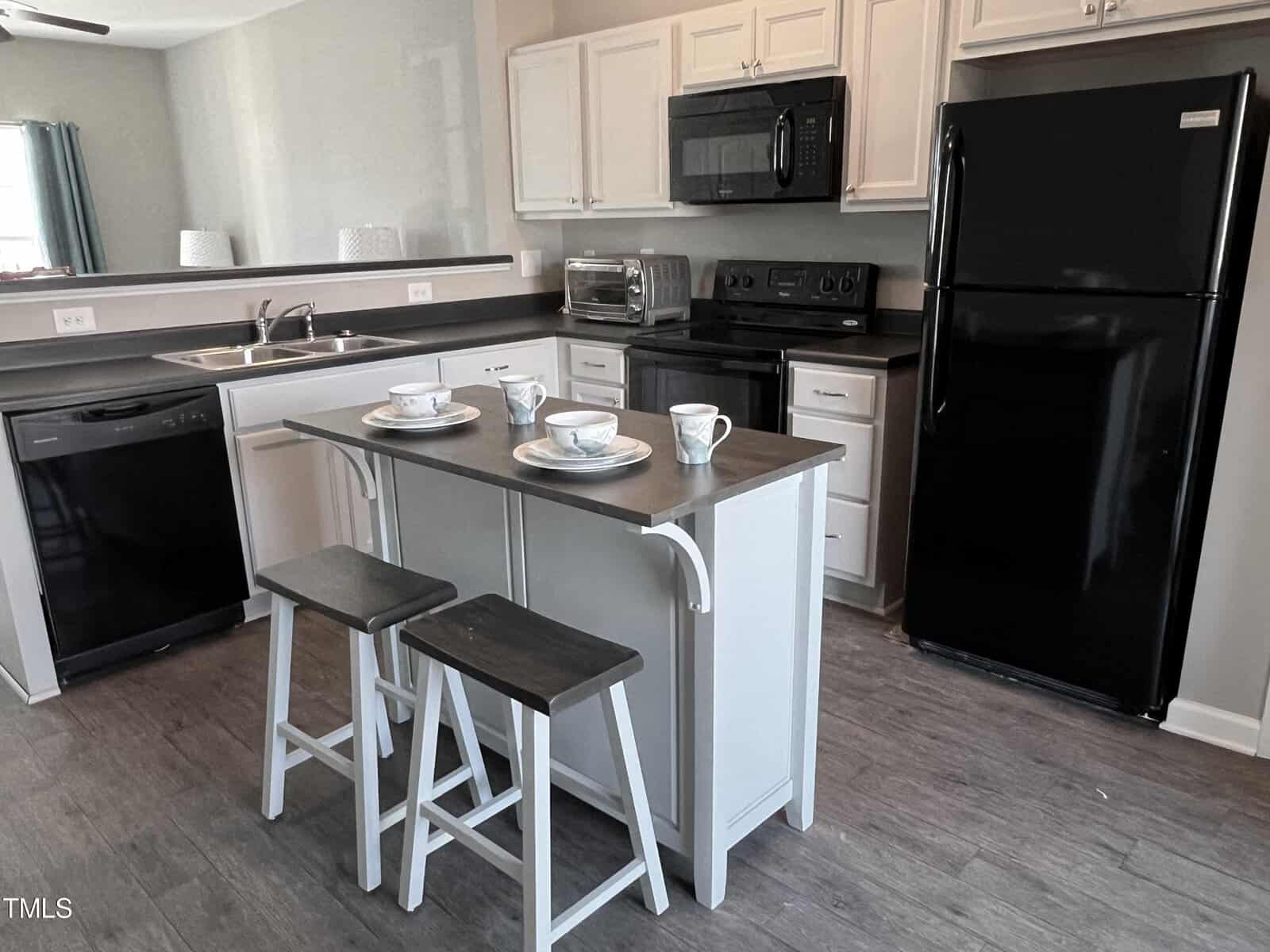 A modern kitchen features white cabinets, a black refrigerator, black stove, and a black dishwasher. A white island with two stools is set with plates and cups. The kitchen has wood flooring and a sink by the window.