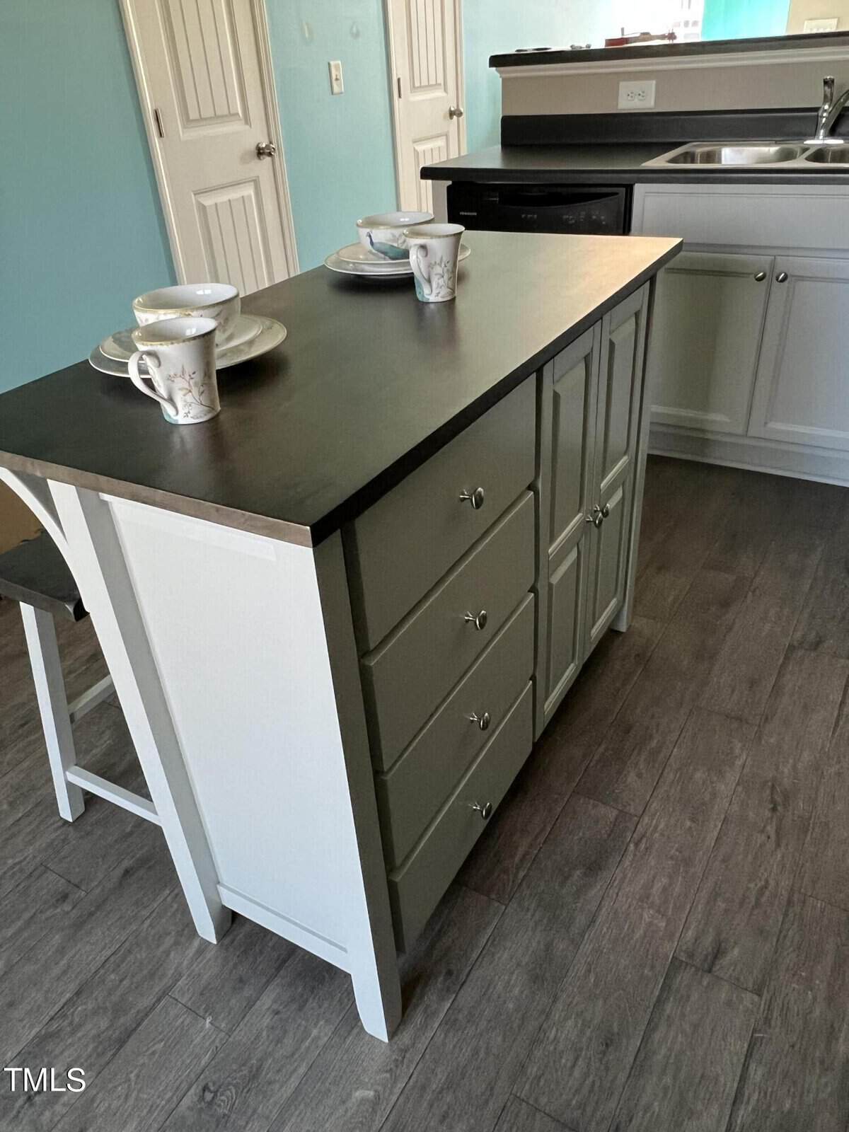 A modern kitchen features a central island with a dark countertop. Two stools are tucked underneath, and white cabinets with silver handles are visible. The island is set for two with cups and saucers. The floor has a wooden appearance.