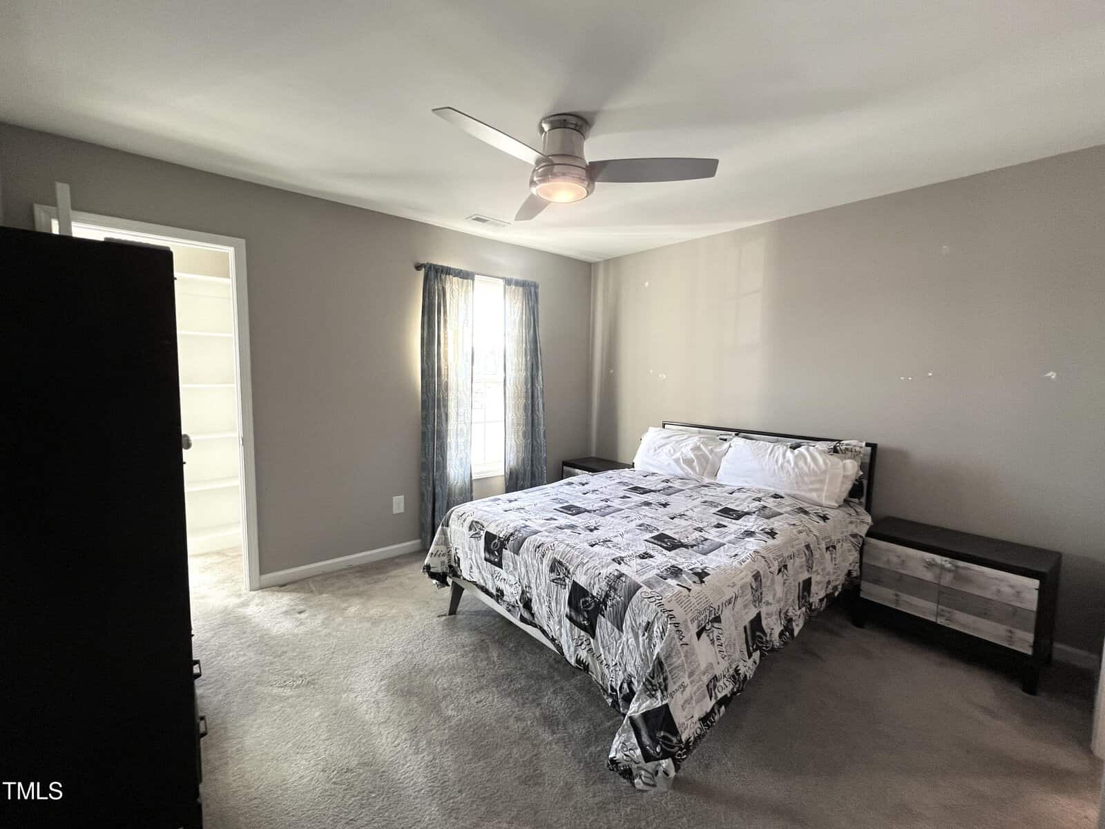 A bedroom with a large bed featuring a black and white patterned comforter. A ceiling fan is above, and there is a window with blue curtains. To the side is a dark wooden dresser, and an open doorway leads to another room.