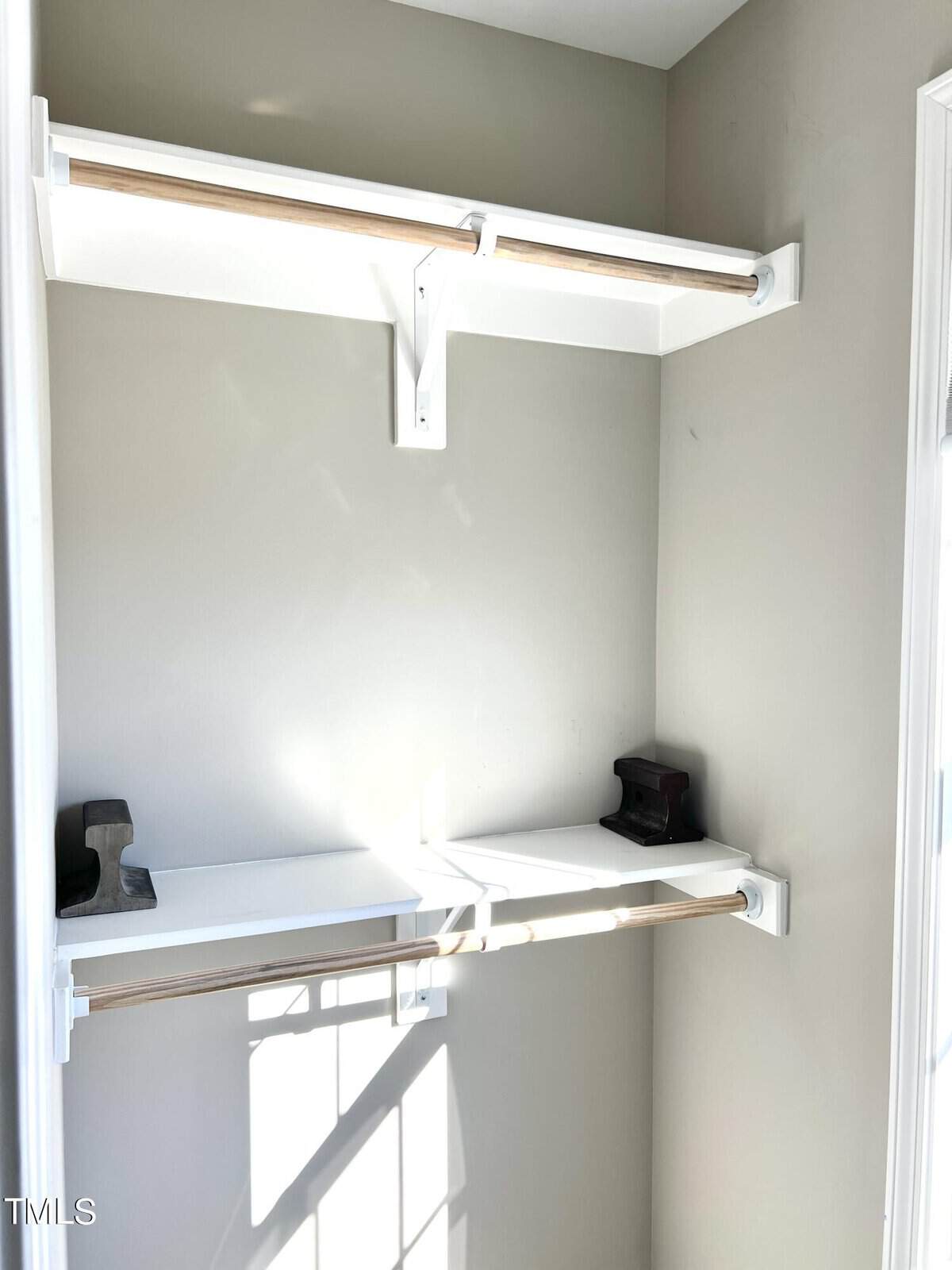 Empty beige closet with two white shelves and hanging rods. Sunlight casts shadows inside.