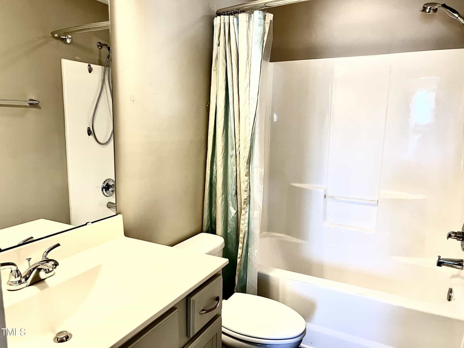 A bathroom with a white sink and counter, a toilet, and a bathtub with a showerhead. A light green shower curtain is partially closed. The walls are painted beige, and a large mirror hangs above the sink.