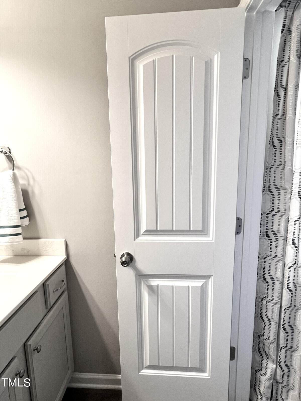 A partly open white bathroom door with vertical paneling leads to a room with gray walls. To the left, a vanity with a white countertop and towel on a rack is visible. A shower curtain with a wavy pattern is on the right.