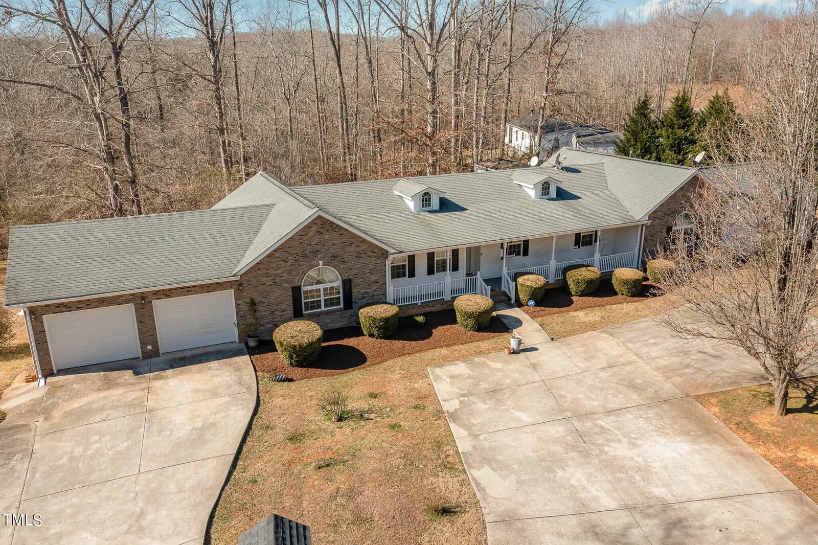 A large single-story brick house with a grey roof and two dormer windows. It has a two-car garage and a covered front porch. Leafless trees surround the property, and a wide driveway leads to the house. The landscape is lightly landscaped.