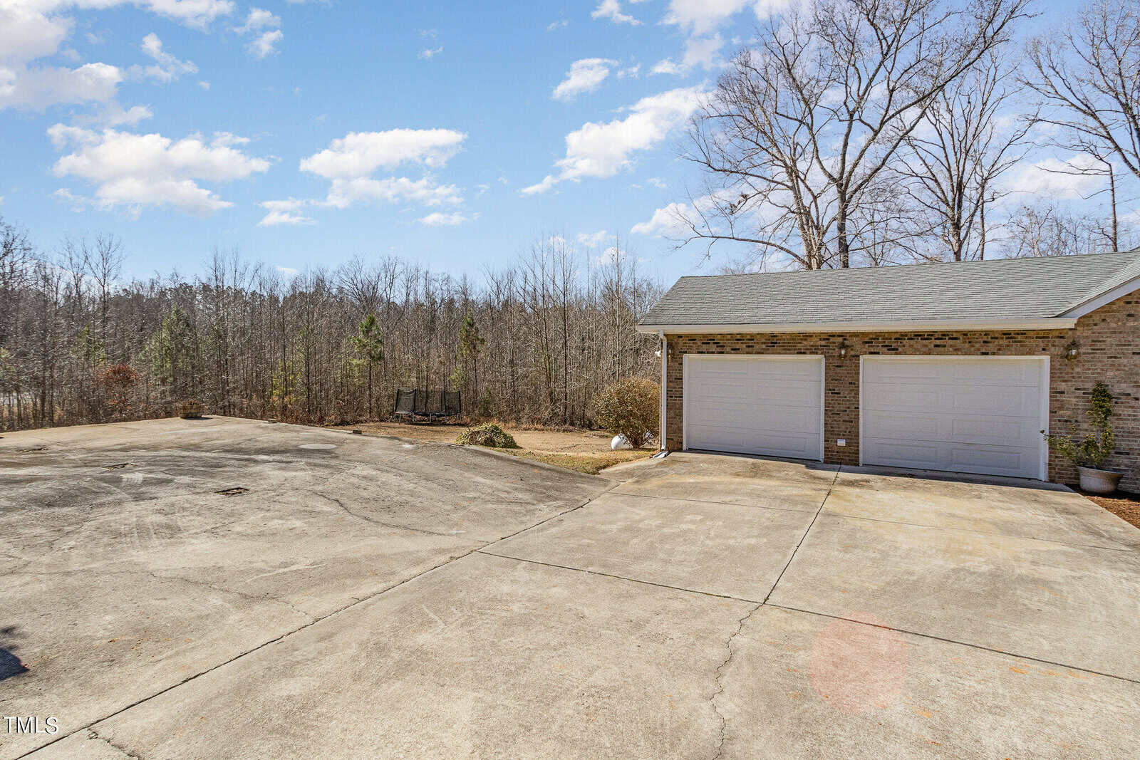 Driveway leading to a two-car garage with white doors attached to a brick building. The area is surrounded by leafless trees under a partly cloudy sky.