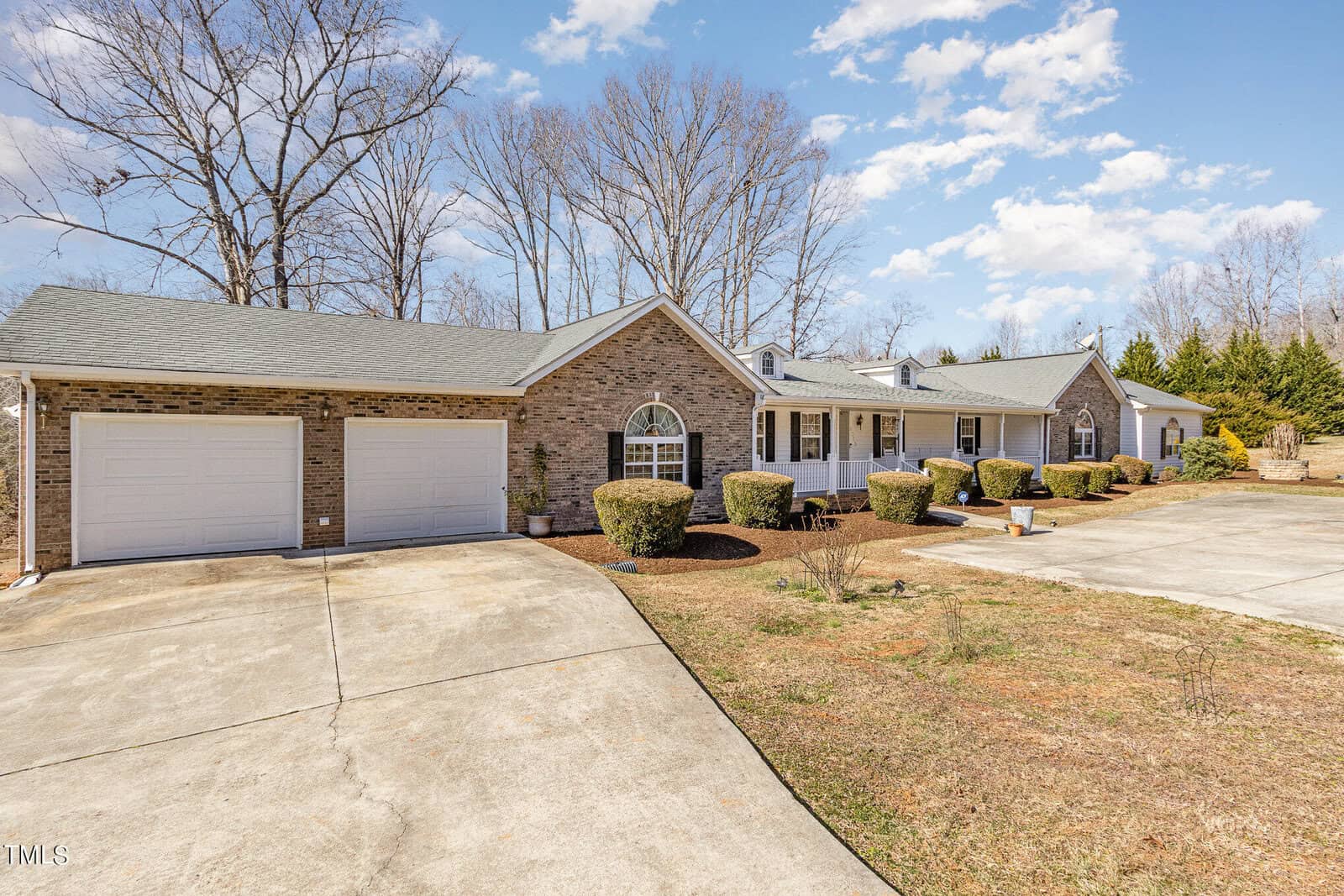 A single-story brick house with a green roof, featuring a two-car garage and a circular driveway. The front yard is landscaped with shrubs and small trees, under a partly cloudy sky.