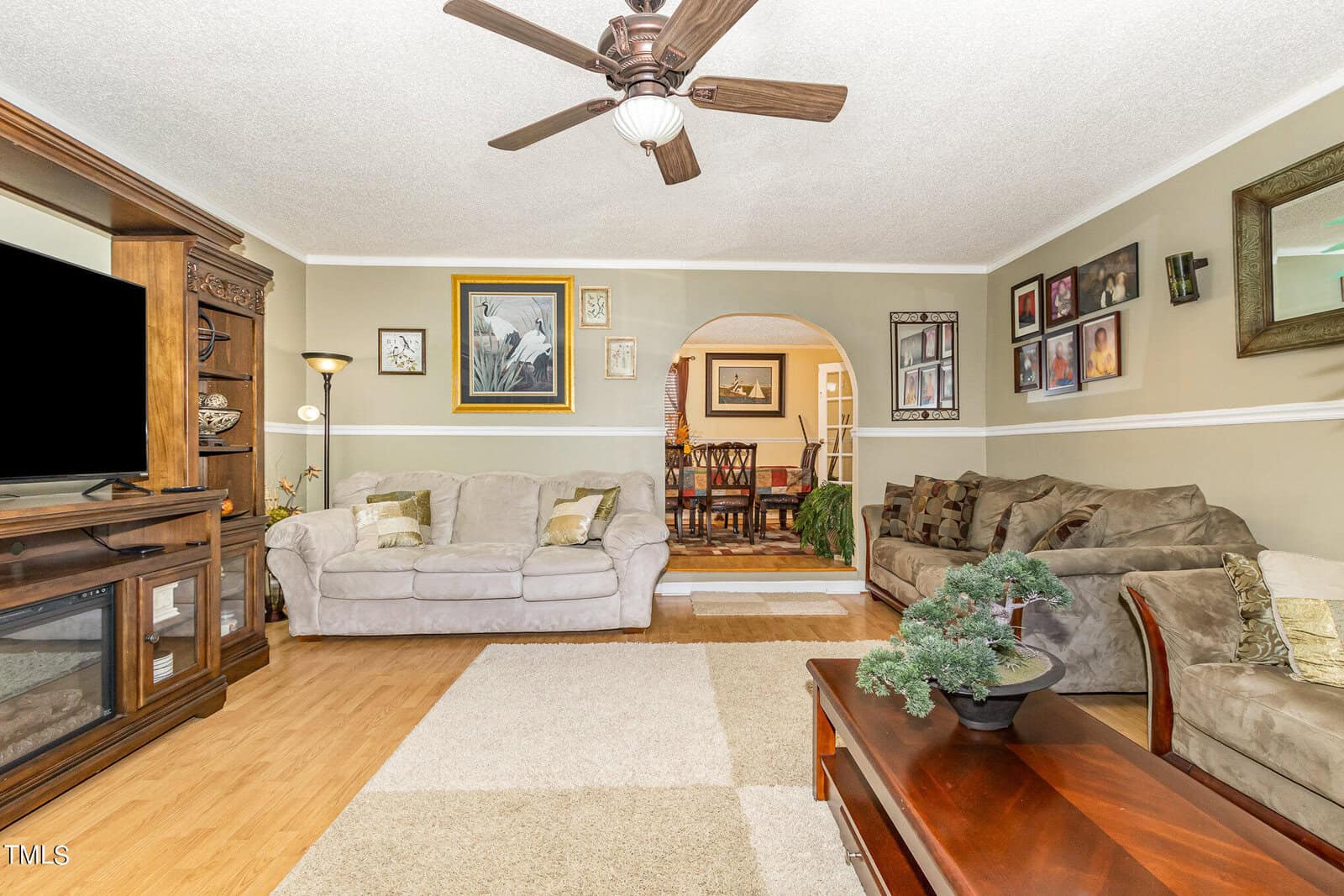 A cozy living room with two sofas, a ceiling fan, and a wooden coffee table adorned with a potted plant. The walls display various framed photos and artworks. An archway leads to a dining room visible in the background.