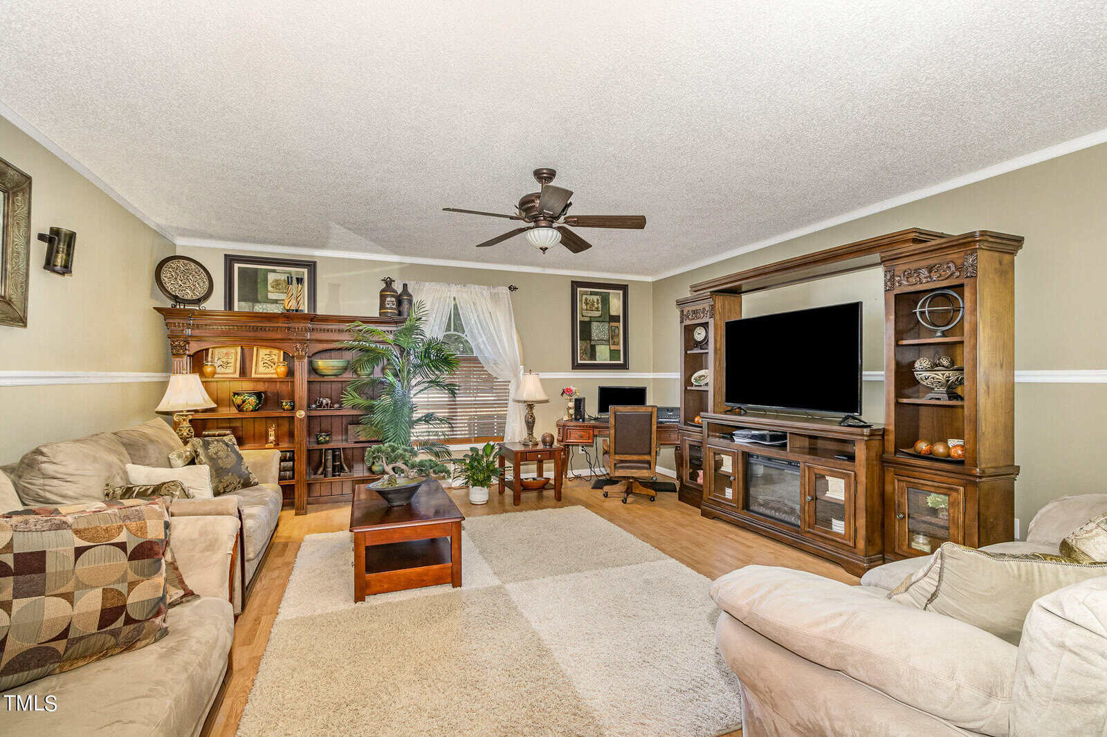 A cozy living room with beige walls features two sofas, a large wooden entertainment center with a TV, and a wooden desk with a chair by the window. Decor includes framed art, a patterned rug, and leafy plants. A ceiling fan is at the center.