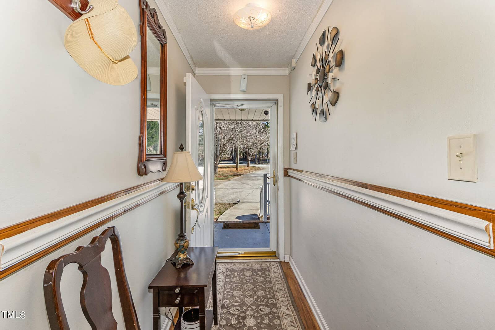 A narrow hallway with white walls and wooden trim leads to an open front door, revealing a view of a yard with trees. The hallway contains a chair, a small table with a lamp, and a hanging hat. A decorative wall clock is visible on the right.