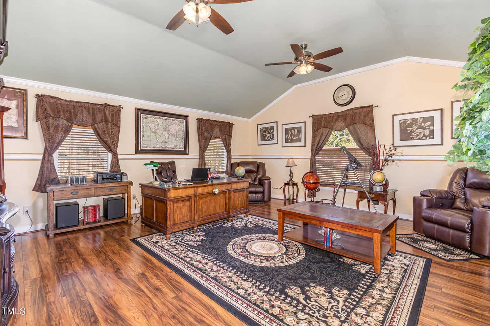 A cozy home office with a wooden desk and leather chairs. Decor includes maps, a telescope, and globes. The room features hardwood floors, large windows with drapes, ceiling fans, and a patterned rug. A potted plant adds greenery.