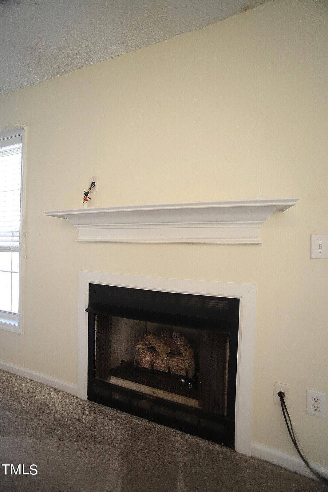 A beige wall with a white framed fireplace below a white mantle. On the mantle, an electrical outlet with exposed wiring is visible. The room is carpeted, and a window with blinds is partially seen on the left.