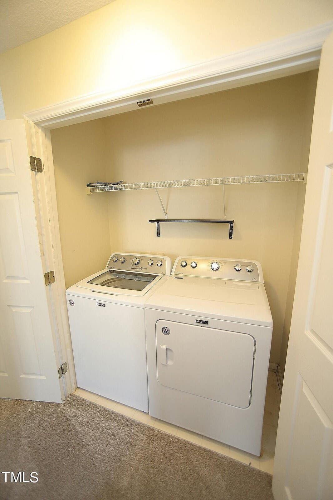 A small laundry nook with a white washing machine and dryer side by side. Above them is a wire shelf and a small black shelf for storage. The area is enclosed by open white folding doors with beige walls surrounding the appliances.