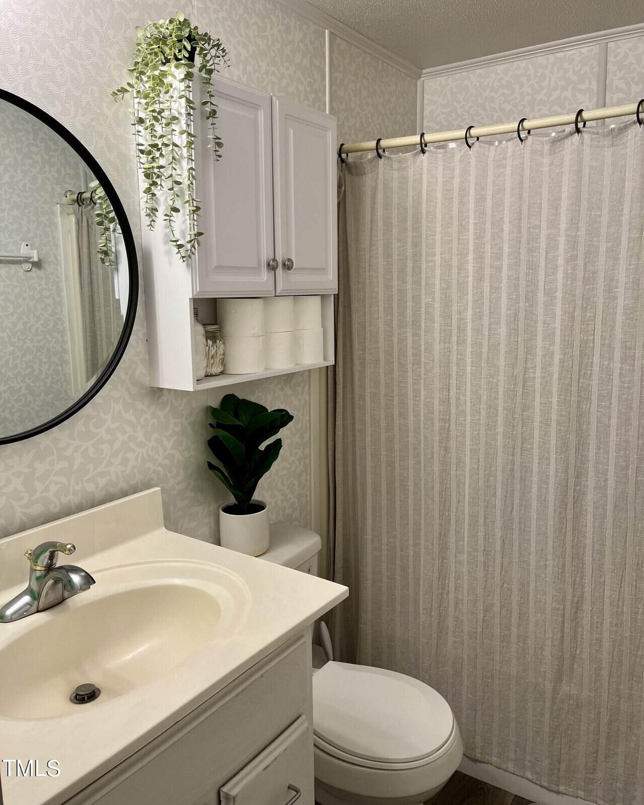 A small bathroom featuring a white vanity with a round mirror, a white sink, and a silver faucet. There's a toilet beside the vanity and a cabinet above it storing toilet paper and a potted plant. A white shower curtain is hung on a rod, and a potted plant decorates the cabinet.
