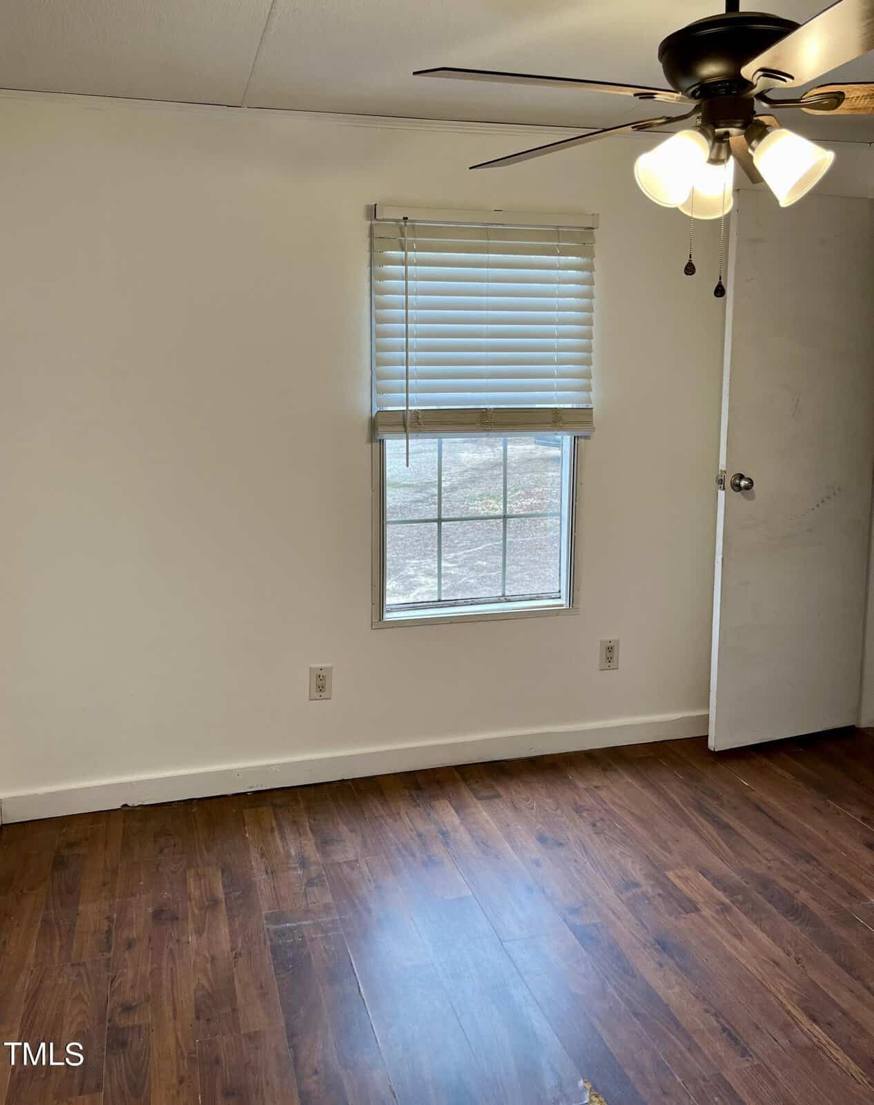 A small, unfurnished room with white walls and wooden flooring. There is a single window with closed blinds, and a ceiling fan with lights above. An open door is partially visible on the right side of the image. The room is well-lit by the natural light coming in through the window.