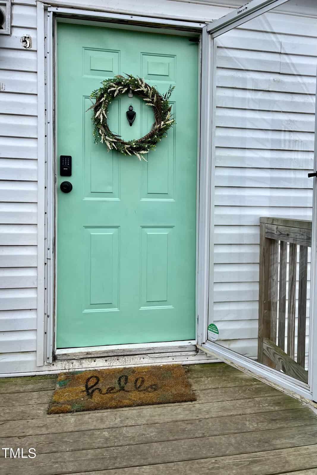 A light green front door at 115 Baileys Creek Drive boasts a wreath made of twigs and greenery. Set in a white paneled house in Kittrell, it features an electronic keypad lock. A doormat with the word "hello" rests on the wooden deck, and a screen door is open to the side.